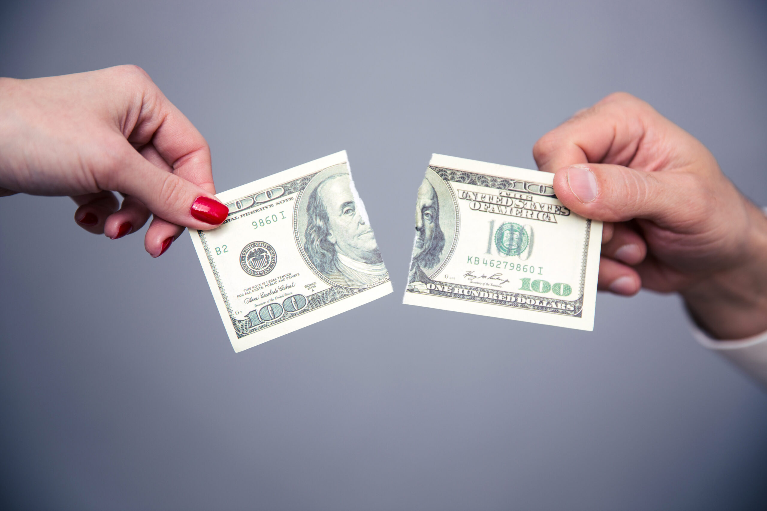 Two hands holding and tearing a hundred-dollar bill, one with red nail polish, against a plain grey background.