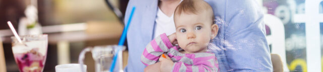 Woman holding a baby in a striped outfit, sitting at a cafe table with desserts and drinks visible in the foreground, waiting for her child visitation attorneys.