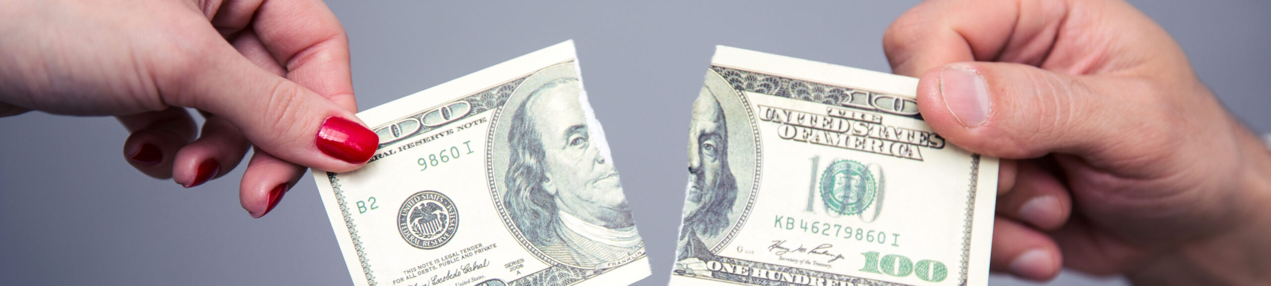 Two hands exchanging a 100-dollar bill during a consultation with property division attorneys, one hand adorned with red nail polish, against a gray background.