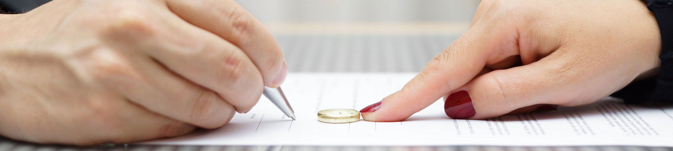 A close-up image of two hands on a document; one hand holds a pen signing the paper while the other hand places a wedding ring beside the signature area.