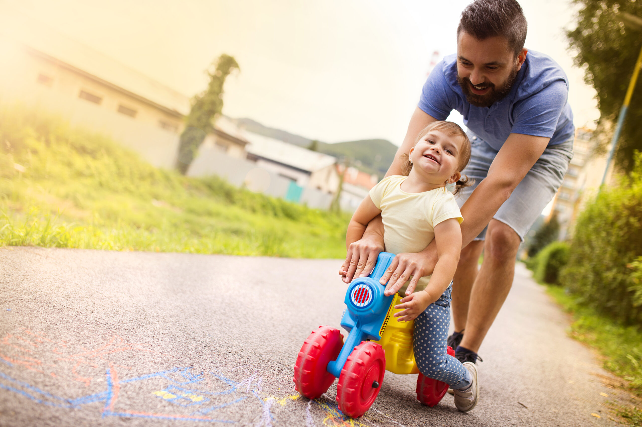 A joyful young child riding a colorful toy truck, guided by a smiling bearded man on a sunlit suburban pathway. the scene is vibrant and full of life.