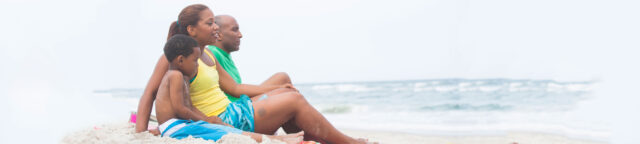 A family sitting closely together on a beach, looking at the sea, with a cloudy sky in the background. They are dressed in summer clothes, radiating relaxation and discussing issues relevant to family law attorneys
