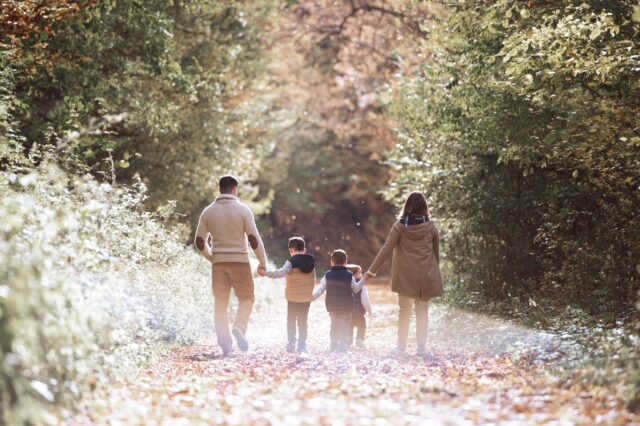 A family of four, with two adults and two children, walks hand-in-hand down a leaf-strewn path surrounded by autumn trees, bathed in sunlight.