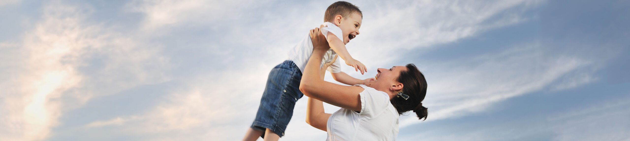 A woman joyfully lifts a young boy into the air against a backdrop of a softly clouded blue sky, both smiling and enjoying the moment, symbolizing the positive outcomes that child support attorneys strive to