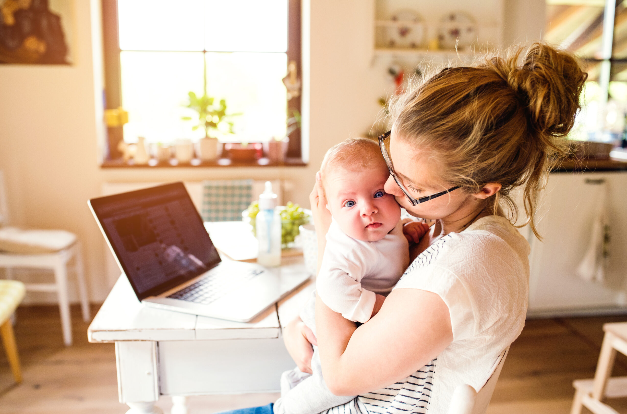 A young woman with glasses, holding a baby, stands near a laptop on a table in a bright, cozy kitchen.