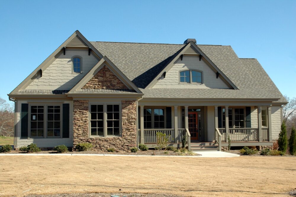 A one-story suburban house featuring mixed siding of grey shingles and stone, a large front porch with columns, and a neatly landscaped yard under a clear blue sky, now on the market due to