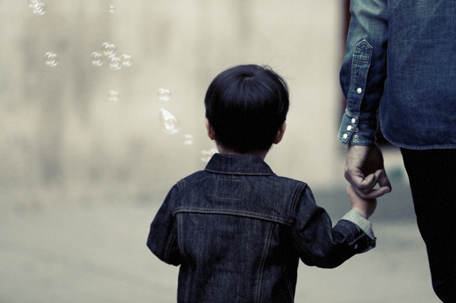 A young child in a denim jacket holds an adult's hand in New Jersey, watching soap bubbles float by in a serene, softly-focused background.