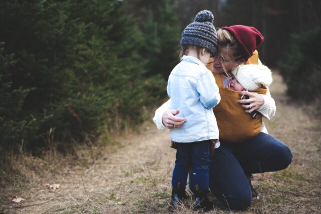 A custodial parent in a yellow jacket and red beanie hugs her two young children, one in a white beanie and the other in a grey beanie, in a forest setting.