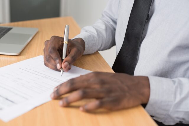 A man in a white shirt and tie signs a prenuptial agreement at an office desk with a laptop nearby. Only his arms and hands are visible.