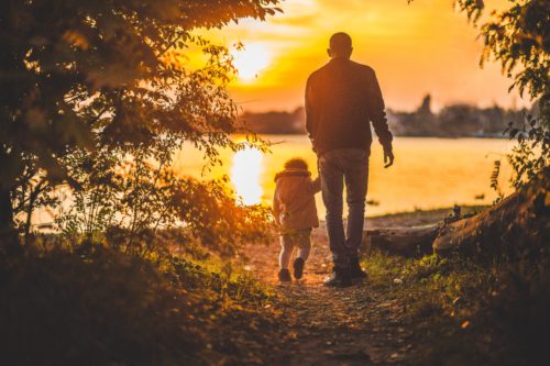 A father and his small child walk hand in hand along a serene lakeside path at sunset, surrounded by trees with sunlight filtering through.