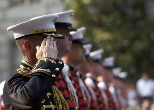 A line of military personnel in ceremonial uniforms saluting, with a focus on one officer in the foreground. They wear white caps and decorated jackets, representing a unit based in New Jersey.