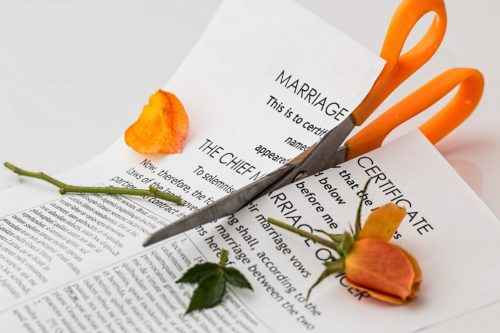 A pair of scissors lies on a marriage certificate, cutting it in half, with a wilted orange rose beside it. This image suggests the concept of divorce or the impact of ending a marriage.