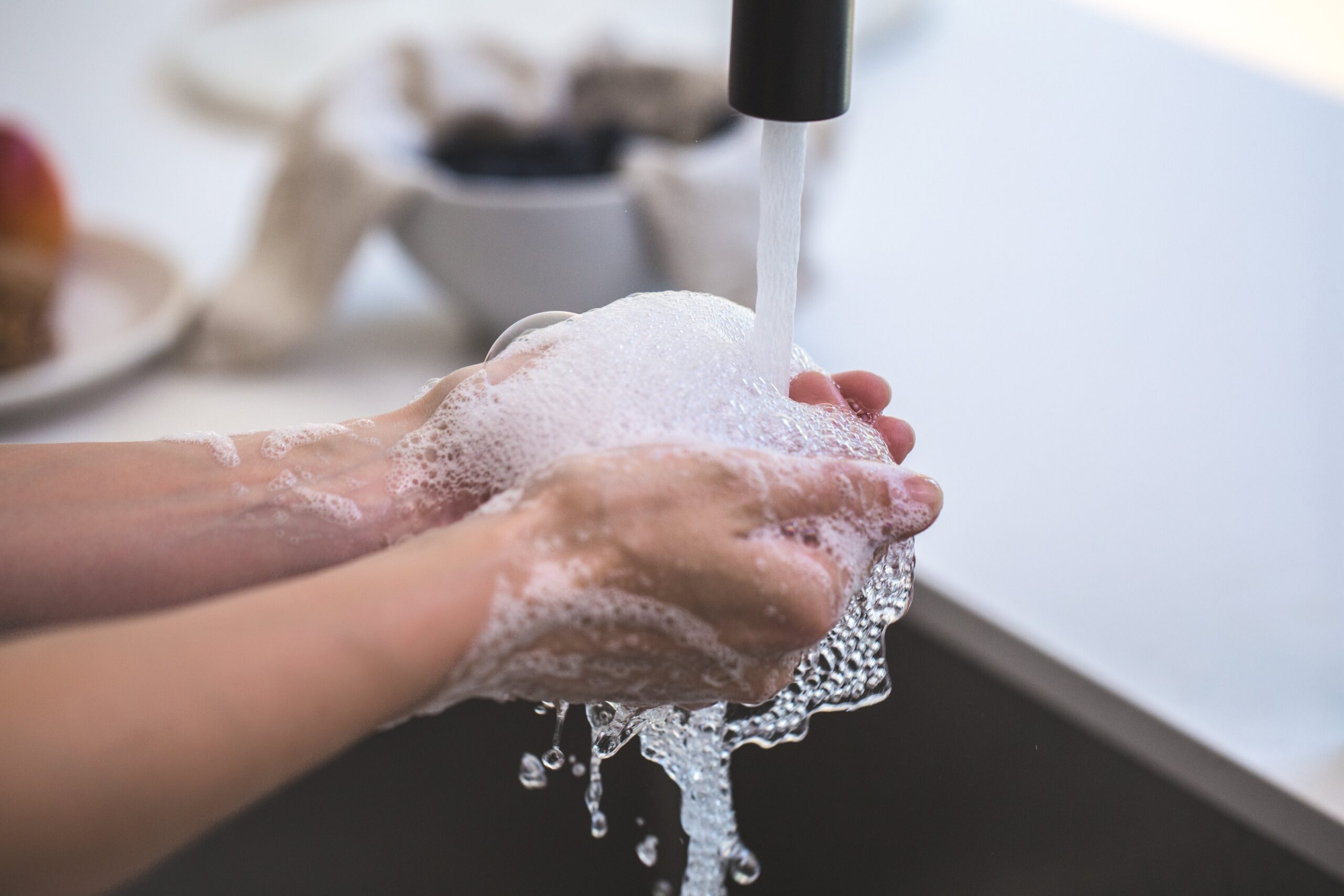 A person washes their hands under a running faucet, lathering with soap suds that drip back into the sink, highlighting cleanliness and hygiene practices essential for preventing COVID-19 transmission.