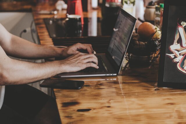 A person is typing on a laptop placed on a wooden table, with a smartphone beside it, avoiding posting on social media. Nearby, a basket containing oranges and an artwork is visible on the left side