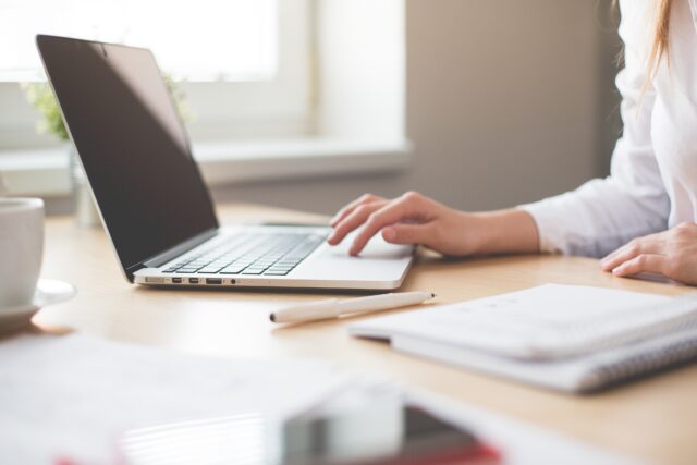 A woman works on a laptop at a desk with papers and a coffee cup, her focus on the screen, arranging an online divorce in a brightly lit room.