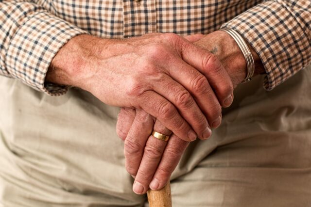 Close-up of an elderly person's hands clasped on a cane, wearing a checkered shirt and khaki trousers, with visible wedding ring and wristwatch, signaling the poignant reality of going through a