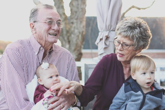 Elderly couple in New Jersey enjoying time with their grandchildren outdoors, the man holding a baby girl and the woman interacting with a toddler boy.