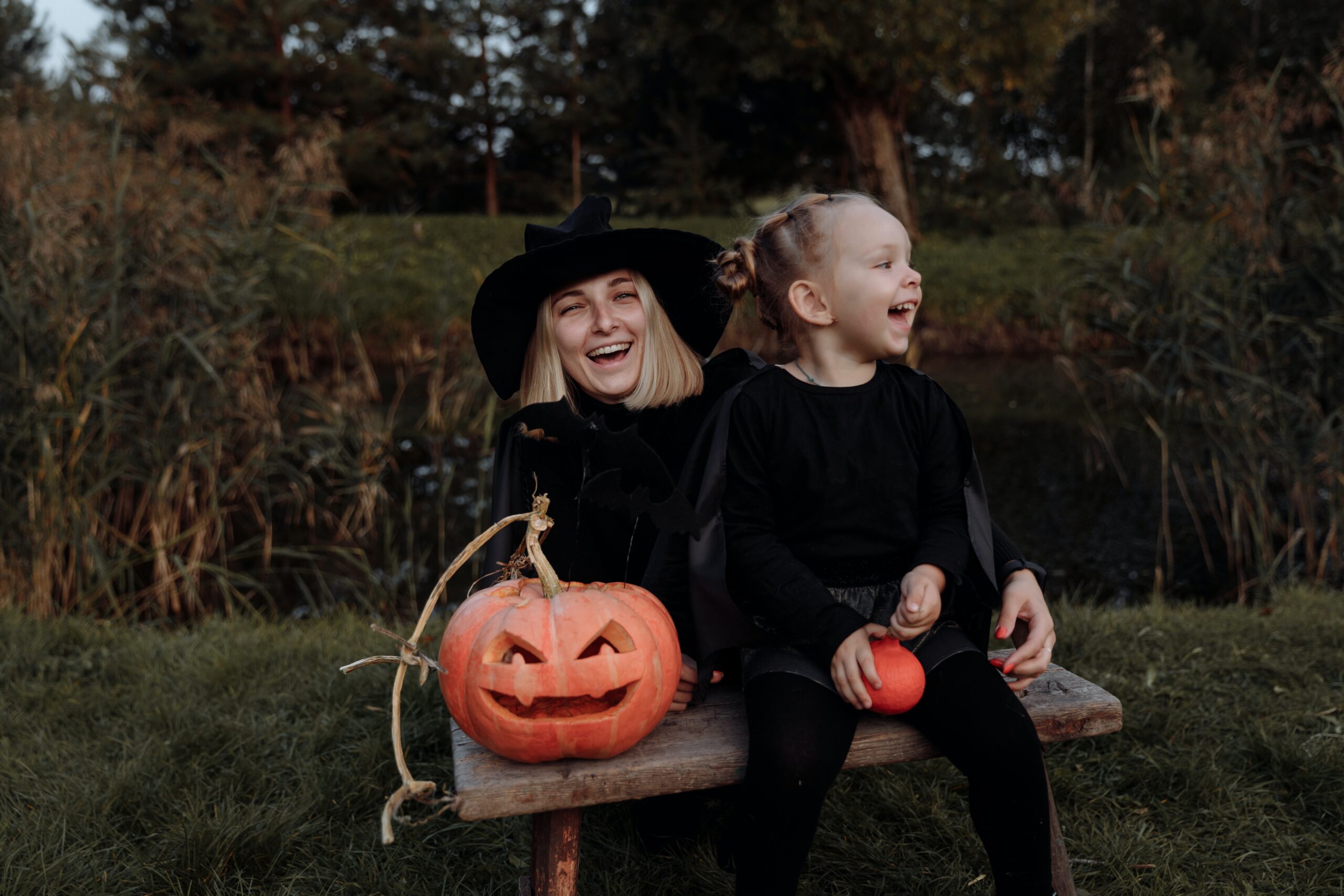 A joyful scene of a young woman and a little girl in black Halloween costumes sitting on a bench outdoors in New Jersey with a carved pumpkin, both laughing heartily.