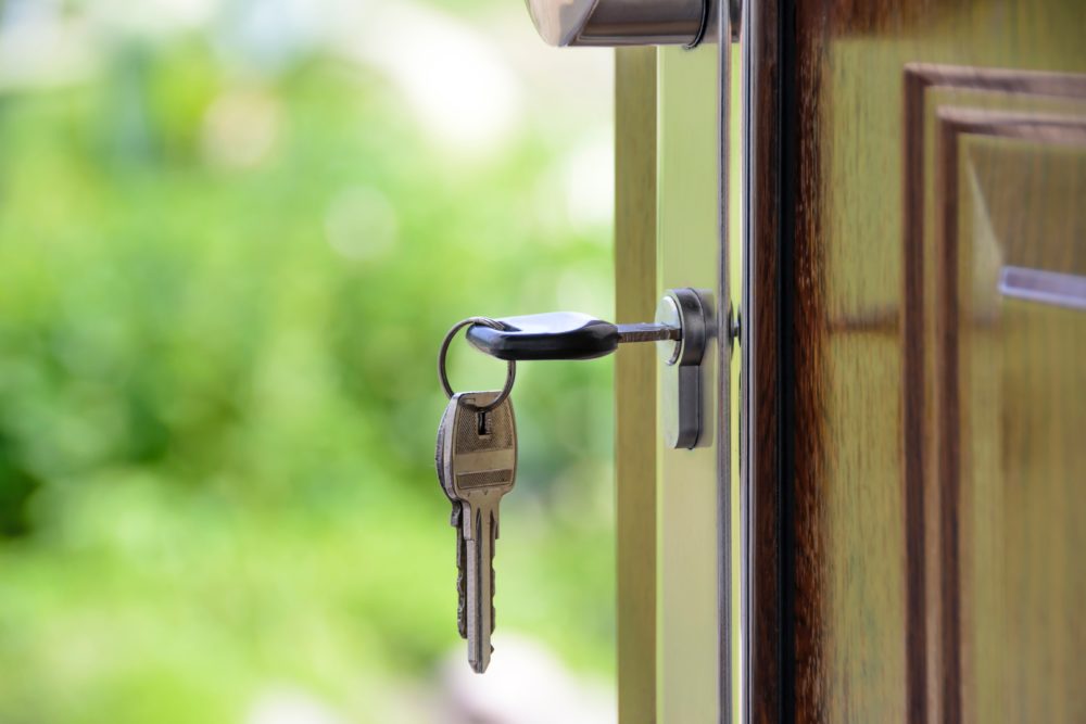 A set of keys hanging from a lock in an open door, with a blurred green background suggesting a garden or outdoor area, symbolizing the question of who gets the house in a divorce.