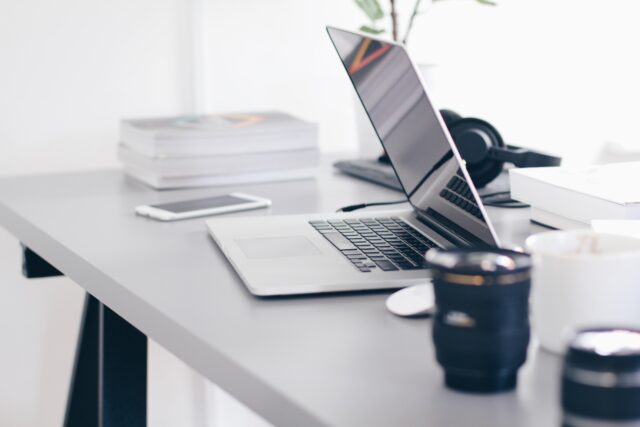 A minimalist workspace featuring an open laptop on a white desk, surrounded by books, a pair of headphones, and a camera lens. The setting has a clean, modern vibe with post-judgment modifications