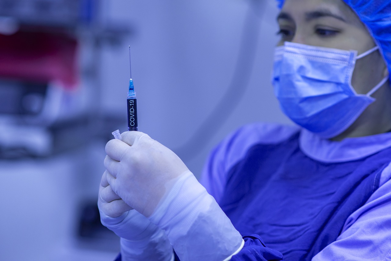A healthcare worker in blue scrubs and a surgical mask holding a covid-19 vaccine syringe, focused on preparing the dose in a clinical setting.