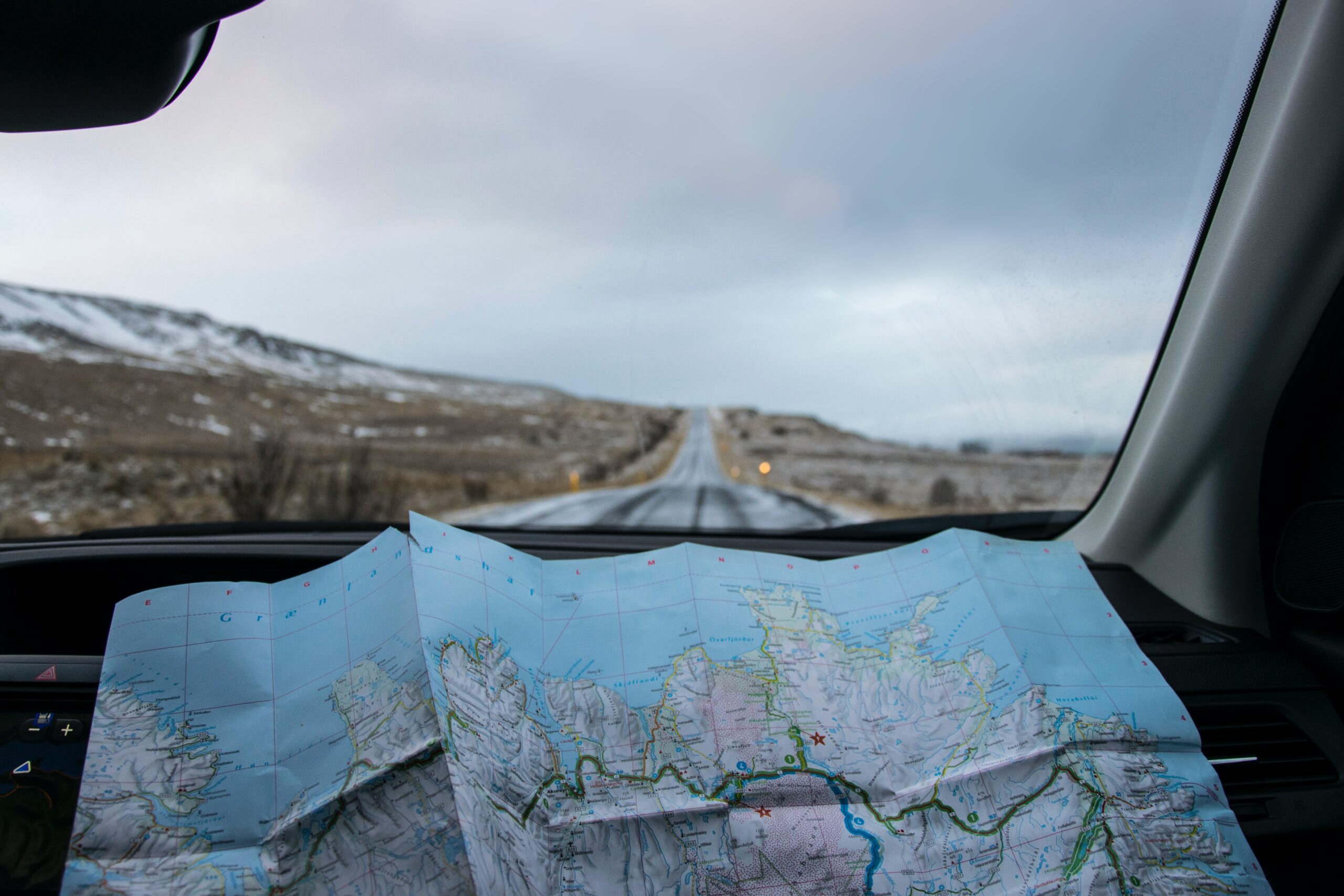 View from a car's passenger seat showing an open road stretching through a snowy landscape, with a map unfolded on the dashboard, suggesting relocation plans.