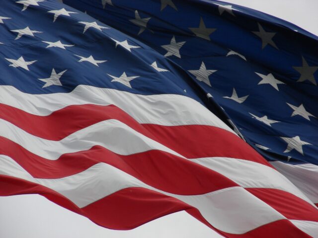 Close-up of the American flag waving, showing the stars and stripes against a grey sky during a military event.
