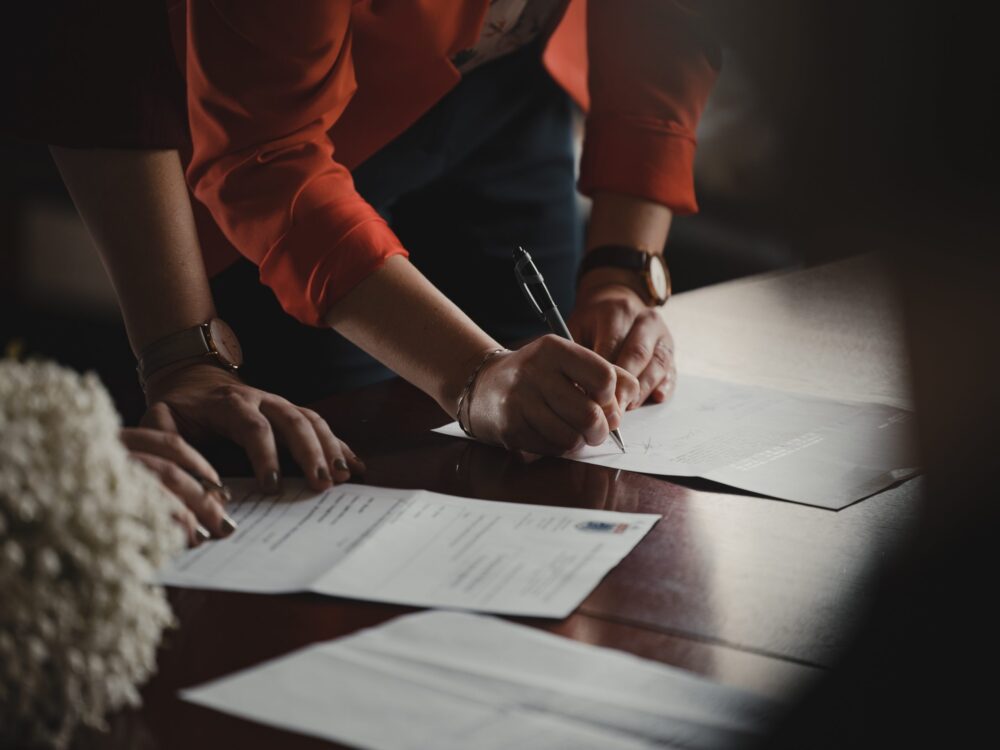 Two people at a table, one wearing an orange shirt, signing a prenuptial agreement with a pen. Focus on the hands and papers, with soft lighting and a bouquet nearby.