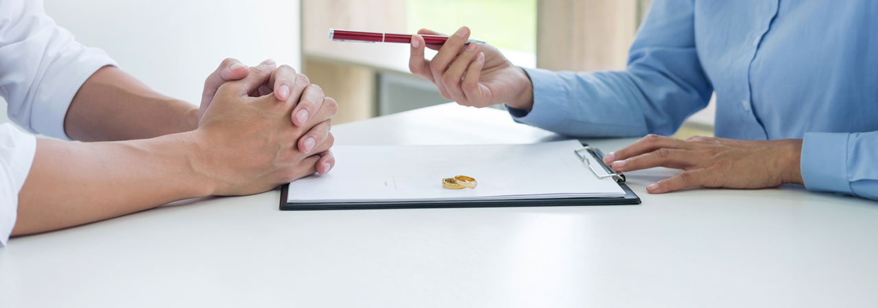 A close-up image of two individuals, seated at a desk, discussing a document where a pair of wedding rings are visible. one person holds a pen, indicating a point on the document.
