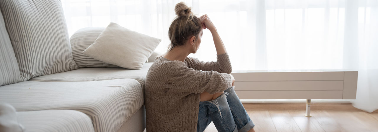 A woman sits on the floor, leaning against a white sofa in a bright room, gazing out of a large window with sheer curtains, her hand resting on her head in a thoughtful pose.