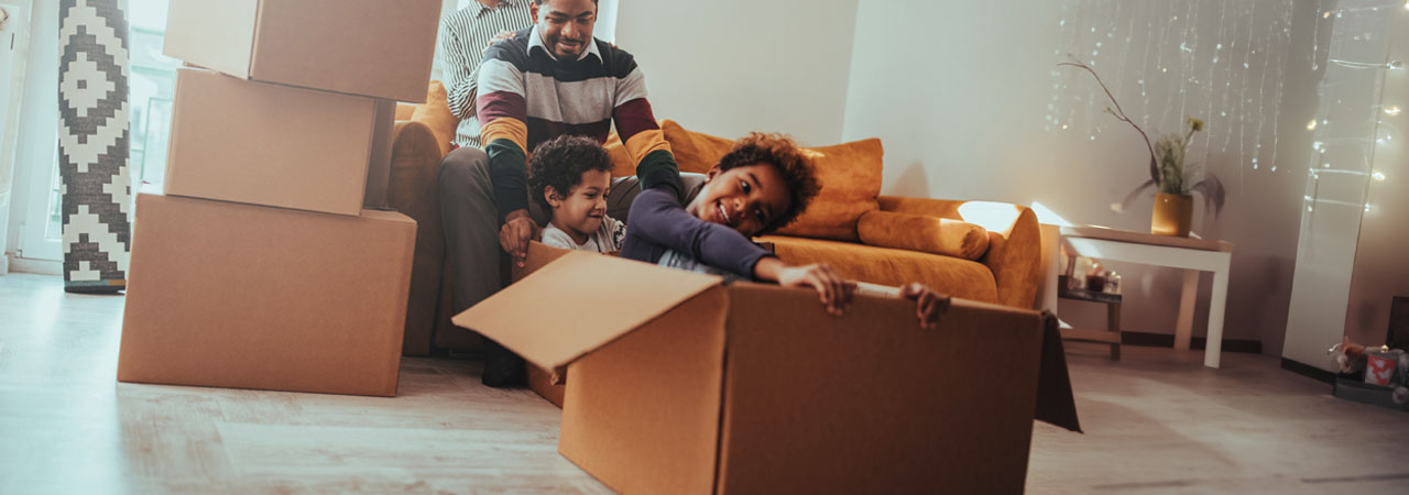 A family enjoys moving day, with a father pushing his child in a cardboard box while the mother watches, all surrounded by unpacked boxes in their new home.