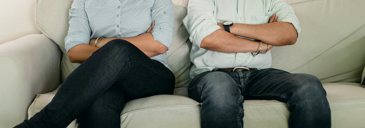 Two people sitting on a sofa with their arms crossed, displaying body language that suggests disagreement or discomfort, focusing on the mid-section and excluding their faces.