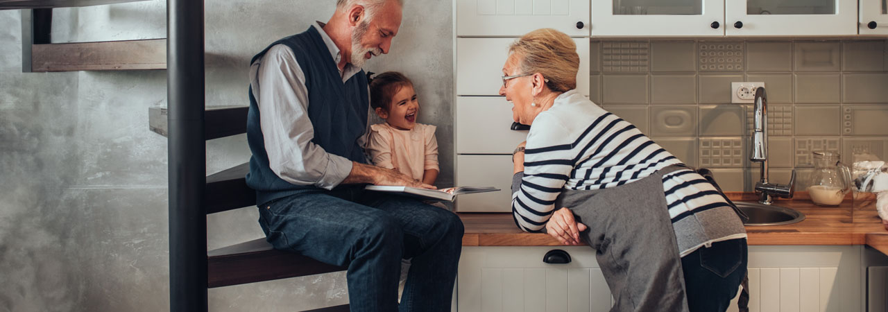 An elderly couple enjoys time with their young granddaughter in a kitchen, sharing laughs and reading a book together.