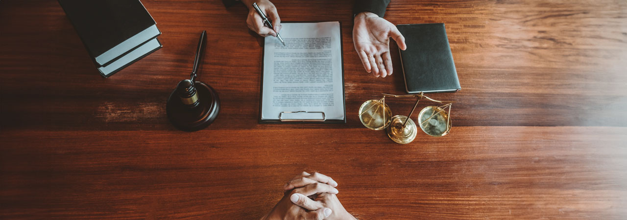Top-down view of a wooden desk with books, a quill in an ink bottle, and spectacles. a person gestures with hands while discussing a document with another person whose hands are visible across the table.