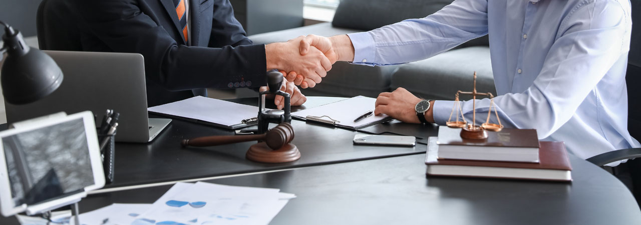 Two individuals shaking hands over a desk in an office, with legal books, documents, and a wooden gavel visible, suggesting a legal or business agreement.