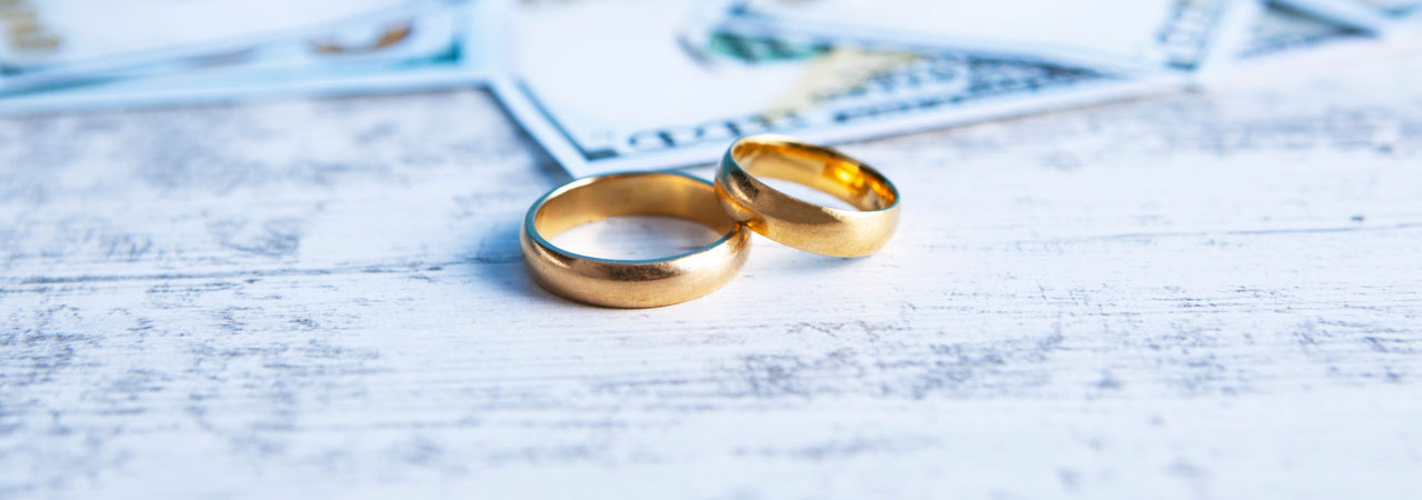 Two golden wedding rings lie on a textured wooden surface, slightly blurred paperwork with financial figures visible in the background.