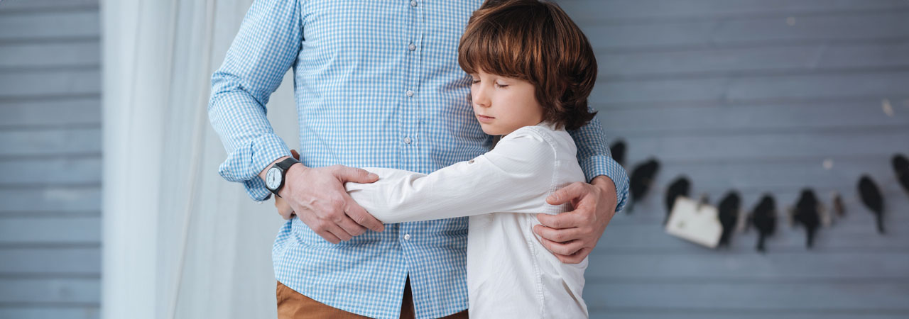 A young boy with brown hair hugs an adult man, seen from the waist down, by a grey wooden wall adorned with small black bird silhouettes.