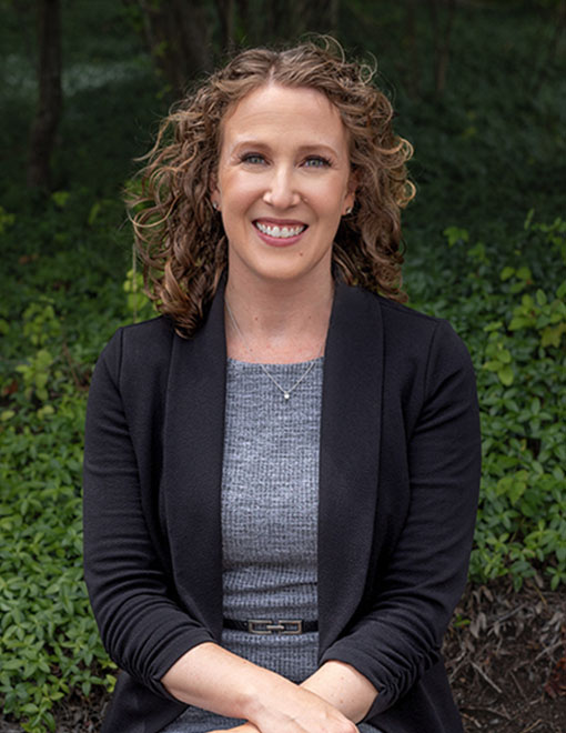 Alison M. Dunn, a woman with curly hair, smiling, wearing a black blazer and grey top, standing before a backdrop of green shrubbery.