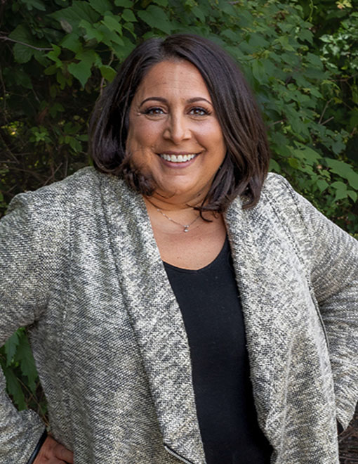 A smiling woman, Jennifer M. Russoniello, Esq., with medium-length dark hair wearing a sparkly silver blazer and black top, stands outdoors with lush greenery in the background