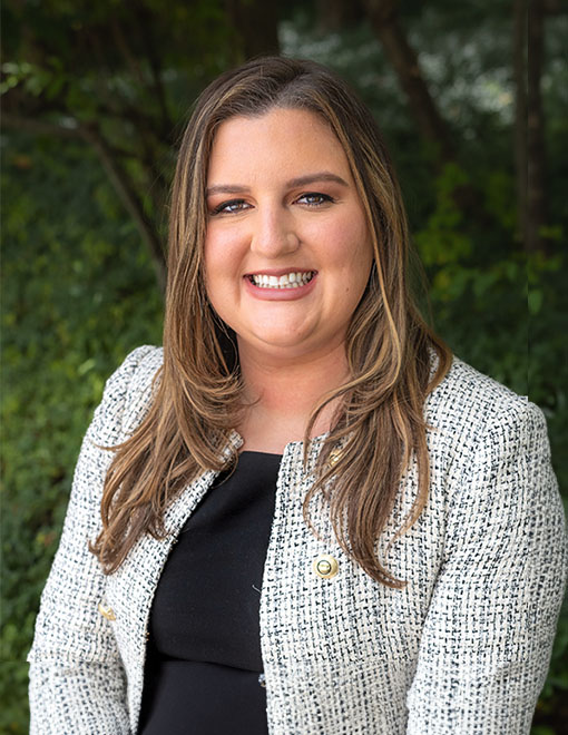 A smiling woman, Lauren E. Sharp, with long, brown hair wearing a tweed blazer and a black top, standing outdoors with greenery in the background.