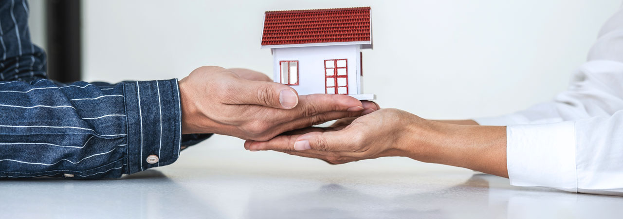 Two people at a table, one handing over a small model house with a red roof to the other, symbolizing a real estate transaction or agreement.