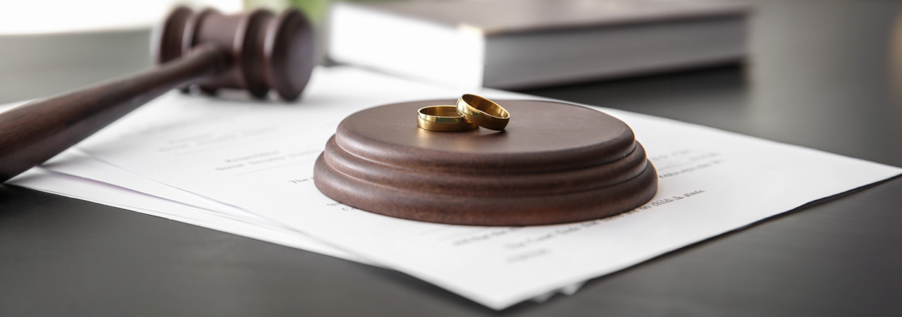 A wooden gavel rests on a desk beside two gold wedding rings, placed overlapping each other on a set of divorce process documents.