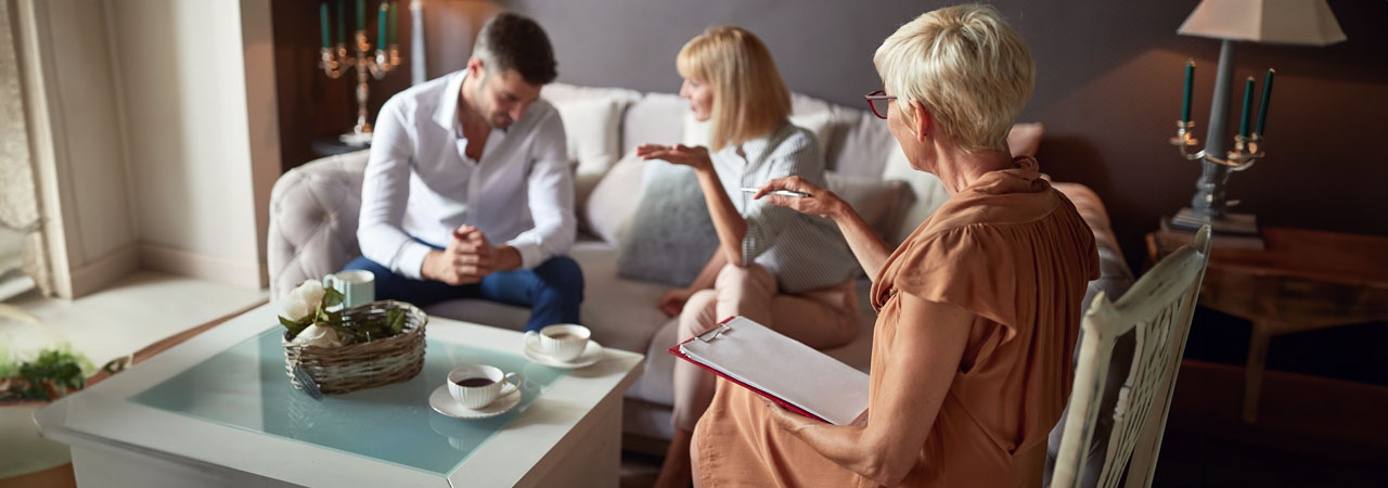 A couple consults with a therapist in a cozy, warmly lit room, sitting on a sofa opposite the therapist who is taking notes. Morris County divorce mediators concept image