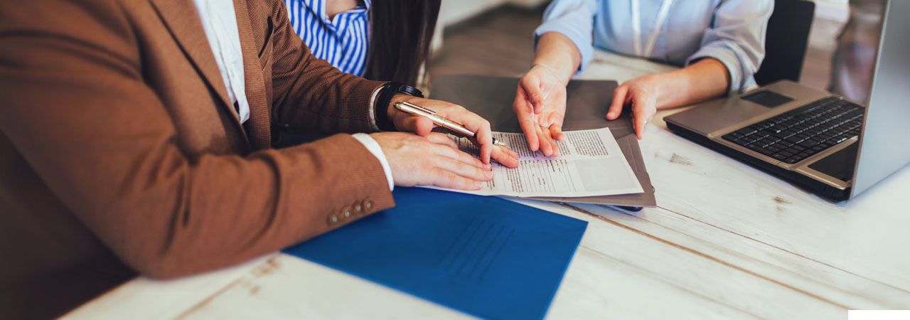 Two professionals seated at a table reviewing a document together, with a laptop and a blue folder on the table. one is pointing at the document while the other holds a pen.