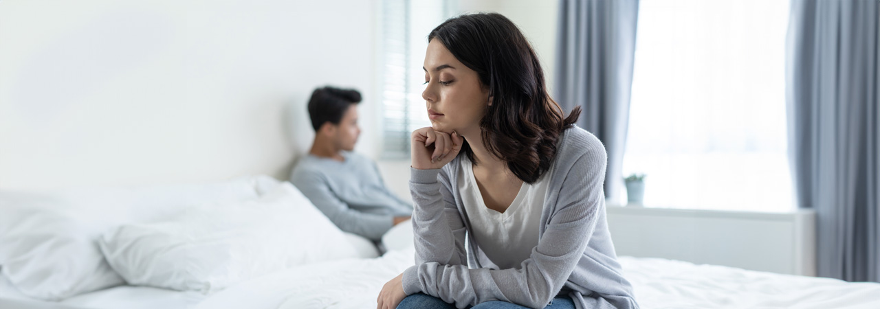 A woman looks thoughtful, sitting on the edge of a bed, with a man sitting in the background, slightly blurred. they appear distant and reflective; Morris County Divorce Lawyer concept image